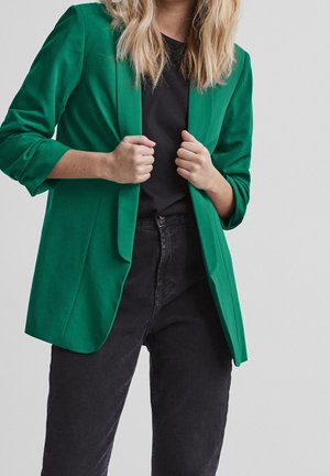 Femme portant un blazer vert sur une chemise noire et un jean noir, tenant les revers du blazer avec les deux mains.
