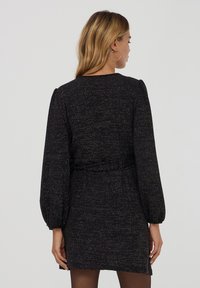 Vestito nero texturizzato con maniche lunghe a sbuffo, vita con cintura e fili metallici leggeri. Scollo semplice rotondo, taglio al ginocchio.