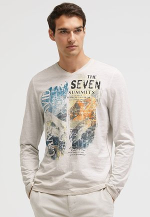 Junger Mann trägt ein helles Langarmshirt mit einer bunten Grafik und dem Text "The Seven Summits", steht vor einem einfarbigen Hintergrund.