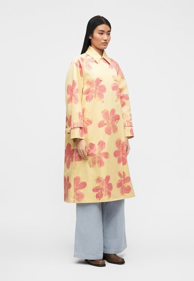 Γυναίκα π ου φοράει κίτρινο διπλής σειράς παλτό με μεγάλα ροζ λουλουδένια σχέδια, φαρδιά ανοιχτό μπλε παντελόνια και καφέ παπούτσια, που στέκεται μπροστά σε λευκό φόντο.