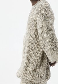 Personne portant un pull en maille beige et blanche avec texture, étendant légèrement le bras gauche vers l'arrière sur un fond clair uni.
