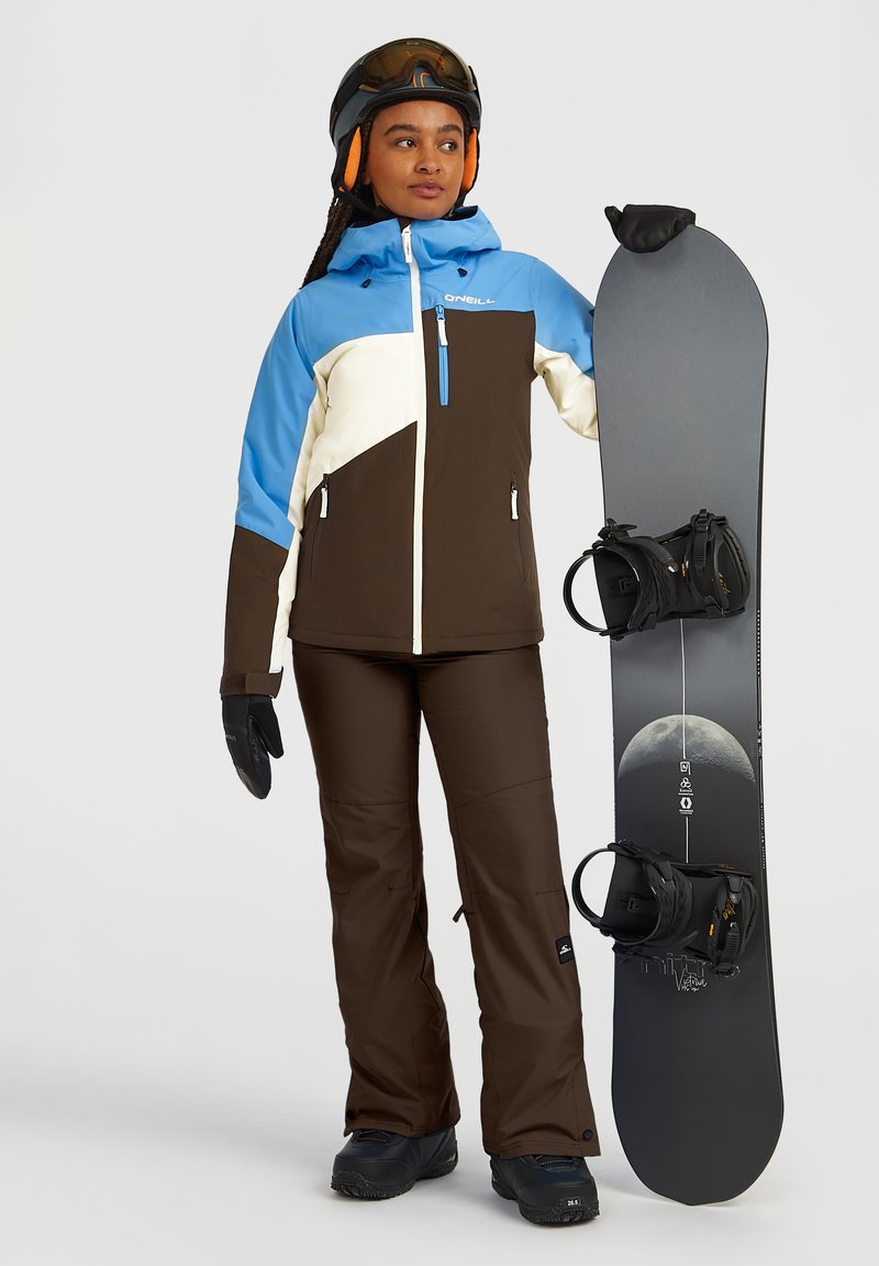 Woman in blue, white, and brown ski jacket and pants holding a black snowboard with moon graphic, wearing helmet and gloves.