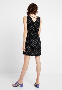 Žena v černých bezrukávových šatech s malými barevnými vzory, se zkříženými ramínky na zádech a fialovými sandály na podpatku, stojící proti bílé zdi.