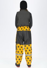 Gele waterdichte broek met een zwart sterrenpatroon, grijze accenten, een elastische tailleband en reflecterende stroken. Gelaagd met een grijze vest en een zwarte top.