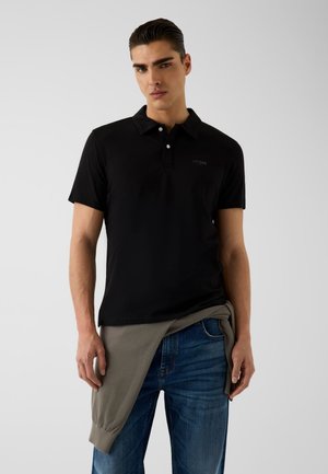 Jeune homme portant un polo noir et un jean bleu avec un pull gris noué autour de la taille, debout devant un fond uni.