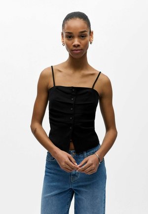 Femme aux cheveux courts portant un haut sans manches noir boutonné et un jean bleu, debout devant un fond blanc uni.