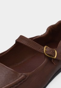Chaussure plate Mary Jane en cuir marron avec une boucle dorée et un détail élastique au talon, vue en gros plan.