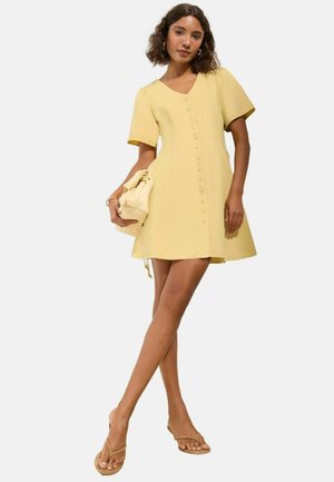 Vrouw die een korte mouw, geknoopte gele jurk draagt, een bijpassende gele handtas vasthoudt en met gekruiste benen in beige sandalen staat.