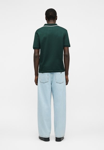 Mężczyzna stojący tyłem, ubrany w ciemnozieloną koszulkę polo z krótkim rękawem i jasnoniebieskie dżinsy o szerokich nogawkach oraz czarne buty na białym tle.