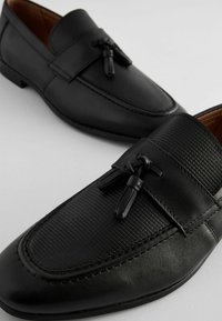 Mocasines negros con cuero liso y acentos texturizados. Presentan un lazo decorativo y detalles de costura. Diseño de punta redondeada y tacón bajo.