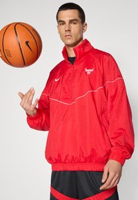 Rød Nike jakke med halvmidte, elastiske manchetter og hvide detaljer. Har et Chicago Bulls-logo på brystet. Sorte shorts med røde striber.