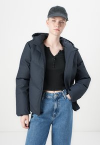 Navy puffer jacket with a hood, featuring a zip closure and ribbed cuffs. Paired with a black henley top and high-waisted light blue jeans.