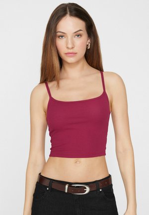 Jeune femme aux cheveux bruns lisses portant un crop top rose foncé ajusté et un jean noir avec une ceinture marron, debout devant un fond uni.