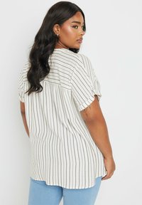 Blusa a righe a maniche corte bianca e beige con vestibilità ampia, dettaglio della piega sul retro e maniche arrotolate, abbinata a jeans azzurri chiari.