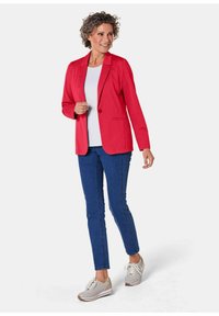 Rode blazer met revers, twee voorzakken, gecombineerd met een witte top en blauwe jeans. Draagt lichte sneakers, model staat voor een neutrale achtergrond.