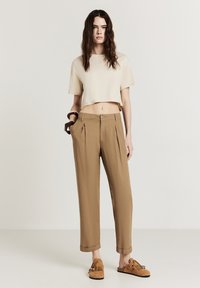 Top corto color crema abbinato a pantaloni plissettati color taupe. I pantaloni presentano tasche laterali e sono traslati in fondo. Calzature marroni con dettaglio frangiato.