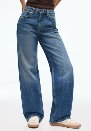 Personne portant un jean en denim bleu taille haute à jambes larges et des baskets blanches, debout les mains dans les poches devant un fond uni.