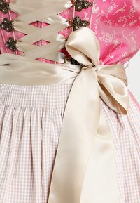 Tenue traditionnelle avec un corsage rose à motif floral, doté de lacets en satin et d'un ruban en satin beige. La jupe présente un motif à carreaux avec une texture froncée.