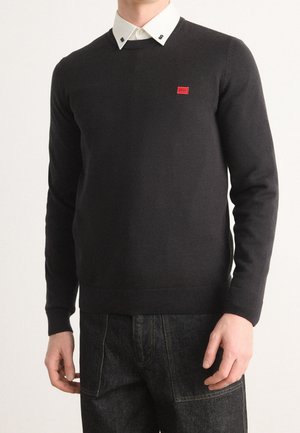 Homme portant un pull col rond noir par-dessus une chemise blanche boutonnée, avec un logo rectangulaire rouge sur la poitrine gauche, associé à un jean foncé.