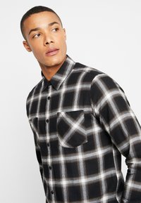Black and white plaid button-up shirt with a pointed collar, long sleeves, and a single chest pocket. Soft fabric texture.