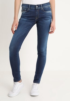 Jeans ajustados de mezclilla azul oscuro, con una cintura de altura media, diseño de cinco bolsillos y un ligero desvanecimiento. Combinados con zapatillas blancas.