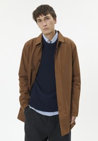 Rjava jakna z dolgimi rokavi z gladko, sijočo teksturo, nošena čez mornarsko moderen pulover in modro srajco z belo črtami, v kombinaciji s temnimi hlačami.