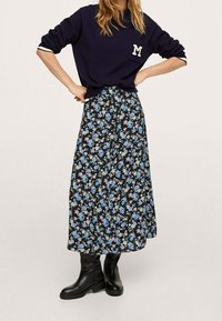 Felpa blu navy con una toppa a forma di "M", abbinata a una lunga gonna nera a fiori con motivi blu, rosa e verdi. Stivali neri al ginocchio.