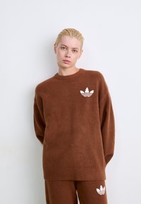 Νεαρό άτομο με κοντά ξανθά μαλλιά φοράει καφέ πλεκτό πουλόβερ και παντελόνι, και τα δύο με λευκό λογότυπο Adidas, στέκεται μπροστά σε λευκό τοίχο.