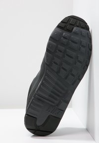 Svart Nike-sneaker sula vinklad mot en vit vägg, med strukturerade mönster och Nike-logotypen nära hälen.