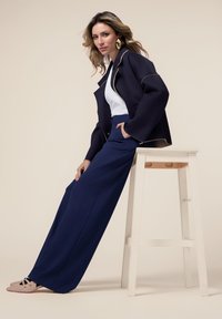 Marina bredvid byxor kombinerade med en vit topp och en marinblå jacka med svarta detaljer. Koordinerande accessoarer inkluderar stora guldörhängen.