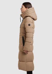 Hosszú, bézs színű puffertakaró magas gallérral és kapucnival. Steppelt mintázat jellemzi, oldalán cipzáras zsebével és logóval ellátott patch-lel a harangon.