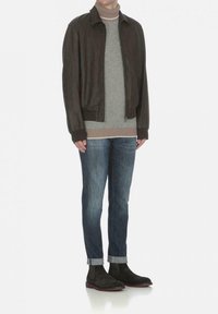 Mörkbrun läderbomberjacka över en ljusgrå stickad polo, kombinerad med blå denimjeans och mörkgröna ankelskor.