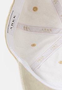 Interior de gorra beige con textura suave, presenta acentos dorados y etiquetas de marca. El forro blanco muestra detalles bordados y costuras.