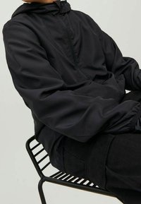 Černá, lehká pulovrová bunda s kapucí, elastickými manžetami a předním zipem. Hladká textura, minimalistický design, nošená přes černé kalhoty.