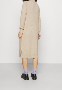 Beżowa dzianinowa sukienka z kołnierzykiem, długimi rękawami i bocznymi rozcięciami, zestawiona z czarnymi butami i lawendowymi skarpetkami. Gładka tekstura i luźny fason.