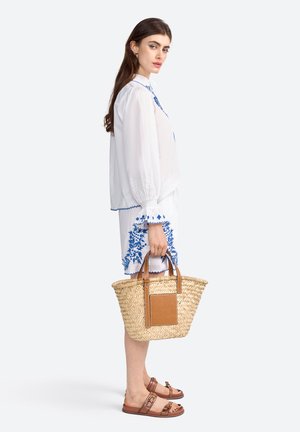 Blouse blanche avec broderie florale bleue, short assorti, sandales en cuir marron et sac en osier avec accent en cuir marron.