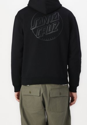 Černá mikina s kapucí s velkým vystouplým logem "SANTA CRUZ" na zádech. Má volný střih a žebrované manžety. Kombinováno s olivově zelenými cargo kalhotami.