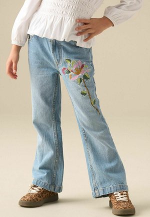 Barn iført lyseblå jeans med en stor pink og grøn blomsterbroderi på højre ben, hvid top og leopardmønstrede sneakers.