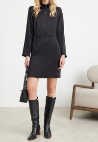 Svart knäppklänning med långa ärmar, figurnära midja, slät textur. Kombineras med knähöga svarta läderstövlar och en liten svart väska.