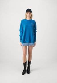 Blauwe sweatshirt met lange mouwen en grafische print, gecombineerd met een versleten denim minirok en zwarte cowboylaarzen. Zwarte pet op het hoofd.