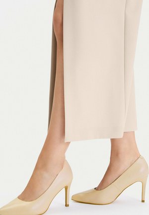 Kobieta w beżowych szpilkach i długiej beżowej spódnicy z bocznym rozcięciem, odsłaniającym część nóg.