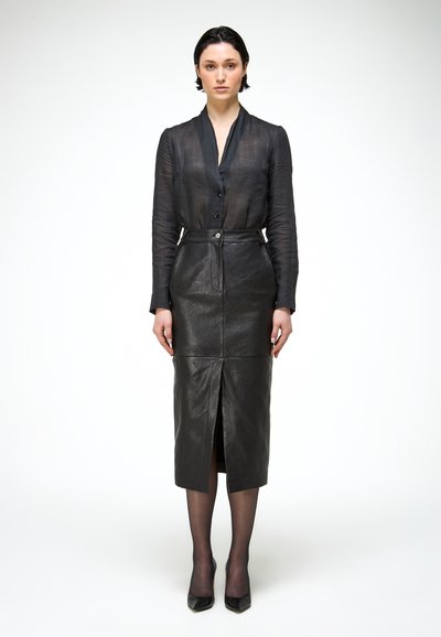 Žena s krátkymi tmavými vlasmi, oblečená v čiernej priehľadnej blúzke, čiernej midi koženej sukni s vysokým pásom a predným rozparkom, čiernych pančuchách a čiernych podpätkoch.