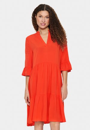 Rochie roșie, până la genunchi, realizată dintr-un material ușor. Are decolteu în V, mâneci trei sferturi cu franjuri și un design cu fuste în straturi.
