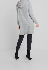 Lång grå cardigan med huva, mjuk textur, ribbade manschetter och sidoflikar, kombinerad med svarta skinny jeans och slanka svarta högklackade stövlar.