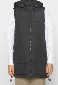 Svart vadderad väst med huva, med dragkedja fram, två sidofickor och justerbar nederkant. Slät textur och minimalistisk design.