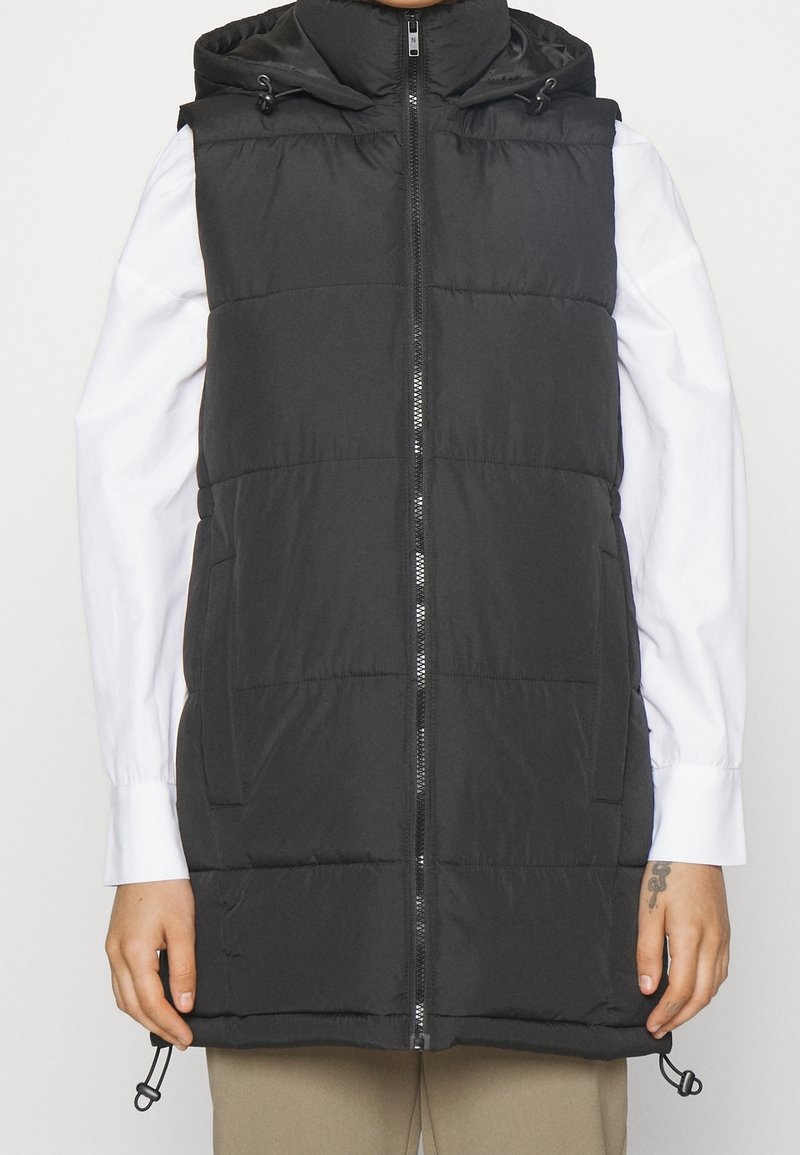 Svart vadderad väst med huva, med dragkedja fram, två sidofickor och justerbar nederkant. Slät textur och minimalistisk design.