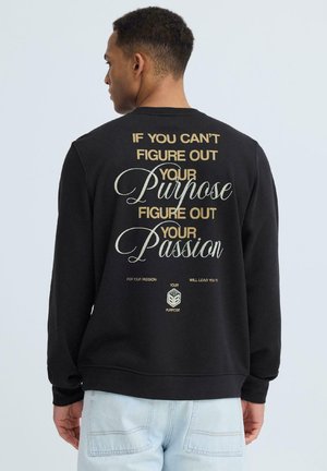 Man wearing black sweatshirt with beige and white text: "If you can't figure out your purpose, figure out your passion." Light blue jeans.