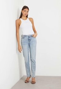 Jean en denim bleu clair avec une légère texture délavée et des détails à motifs, assorti d'un haut blanc sans manches et de sandales à talons noires.