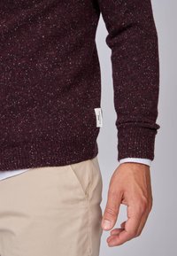 Pull en maille bordeaux avec une texture mouchetée, poignets côtelés et une petite étiquette de marque sur le côté, associé à un pantalon beige.