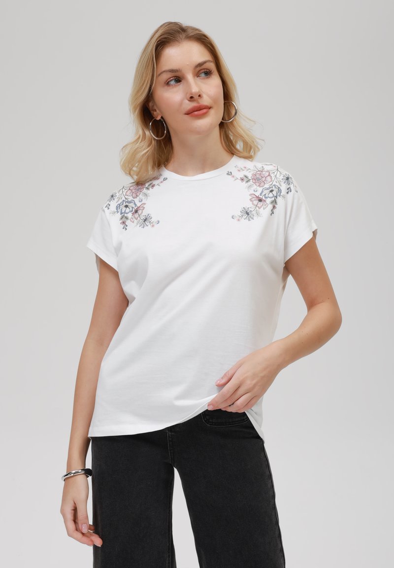 T-shirt en coton à manches courtes de couleur blanche, orné de broderies florales en rose et bleu sur les épaules. Texture lisse, coupe décontractée.